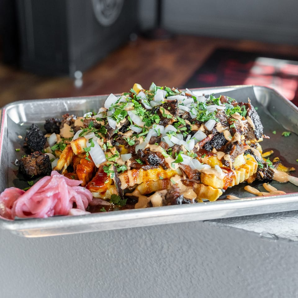 Loaded fries with brisket, onions, cilantro, and sauce, served with pickled onions and a beer.