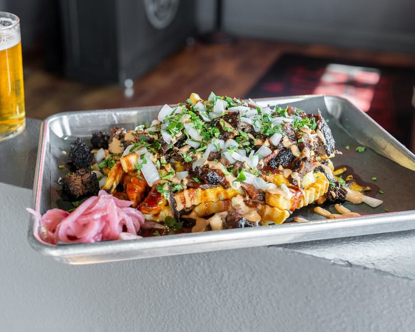 Loaded fries with brisket, onions, cilantro, and sauce, served with pickled onions and a beer.