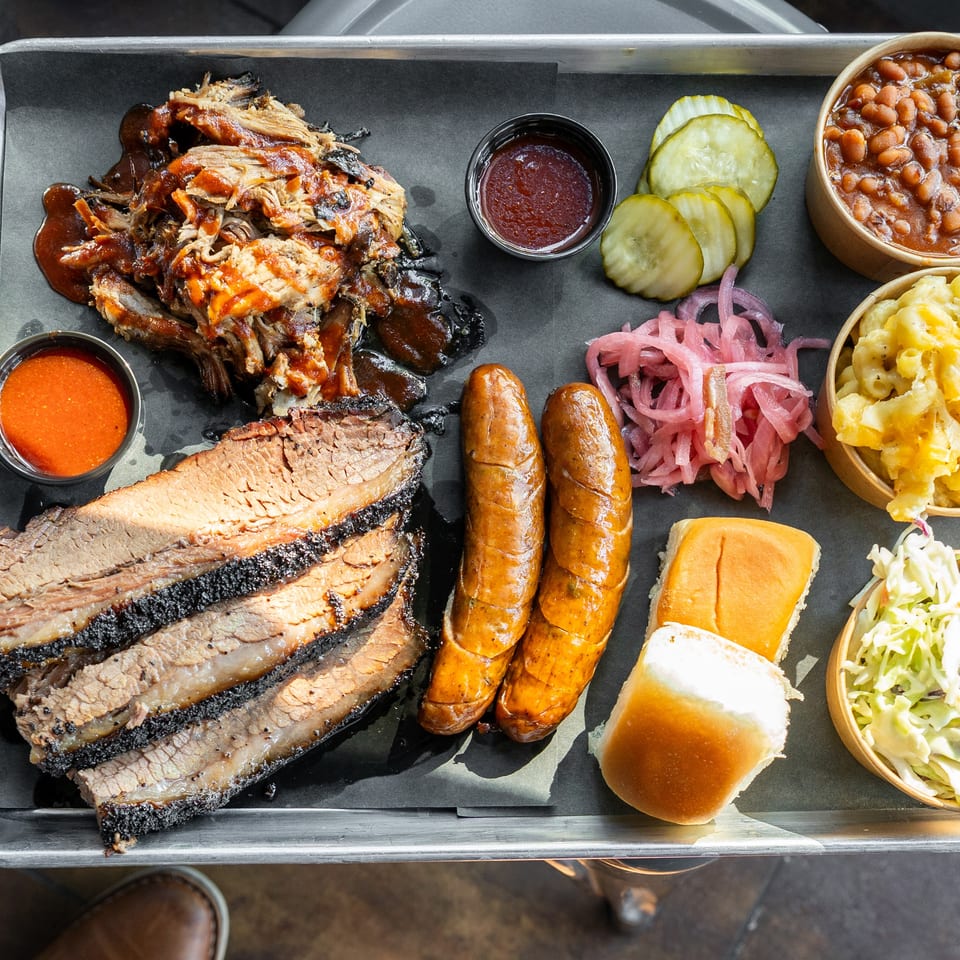 A metal tray with brisket, pulled pork, sausages, baked beans, mac and cheese, coleslaw, and pickles.