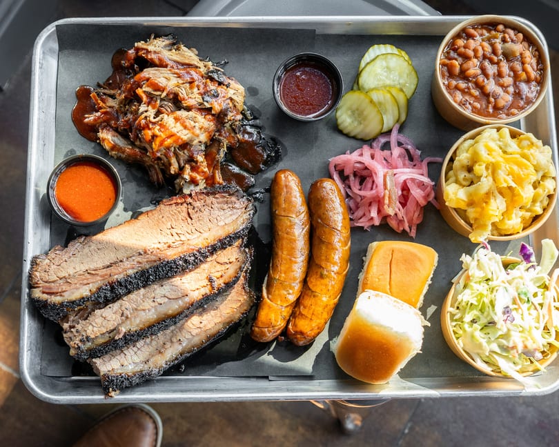 A metal tray with brisket, pulled pork, sausages, baked beans, mac and cheese, coleslaw, and pickles.