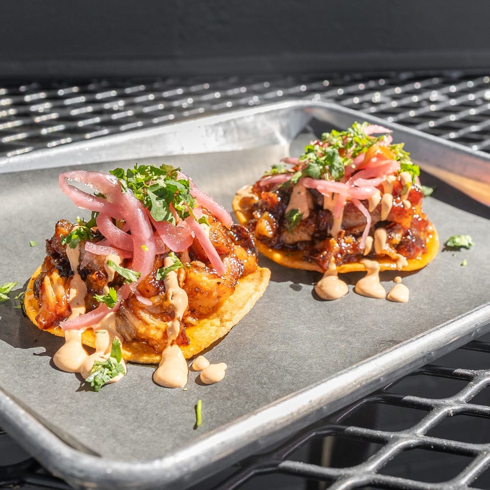 Two gourmet tacos with meat, pickled onions, cilantro, and sauce on a metal tray.