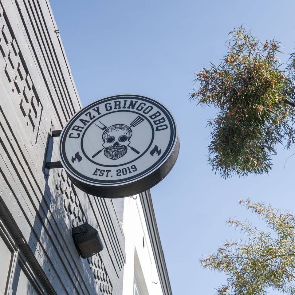 Crazy Gringo BBQ sign with a skull logo, mounted on a gray building under a clear sky.