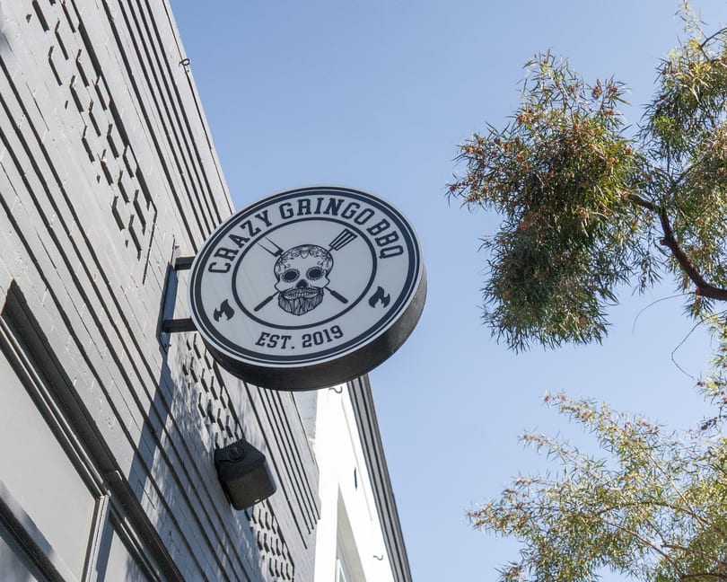 Crazy Gringo BBQ sign with a skull logo, mounted on a gray building under a clear sky.