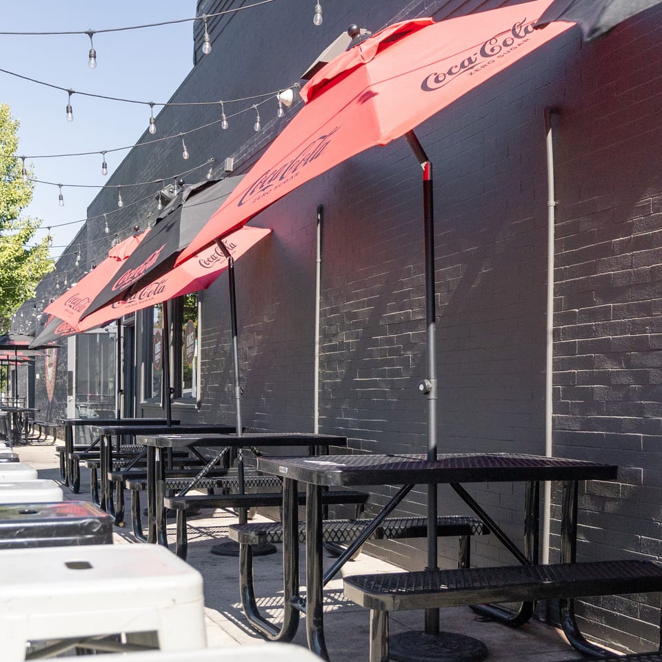 Outdoor patio seating with black picnic tables, red Coca-Cola umbrellas, and string lights against a dark brick wall.
