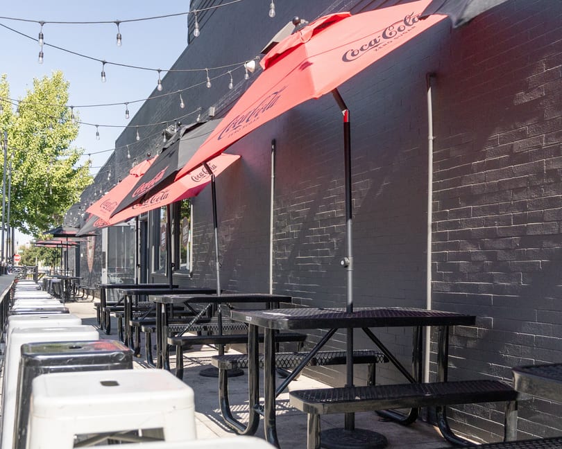 Outdoor patio seating with black picnic tables, red Coca-Cola umbrellas, and string lights against a dark brick wall.