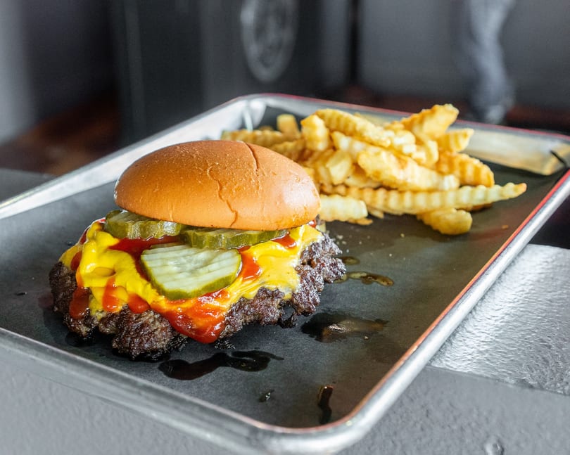 A cheeseburger with pickles, mustard, and ketchup, served with crinkle-cut fries on a metal tray.
