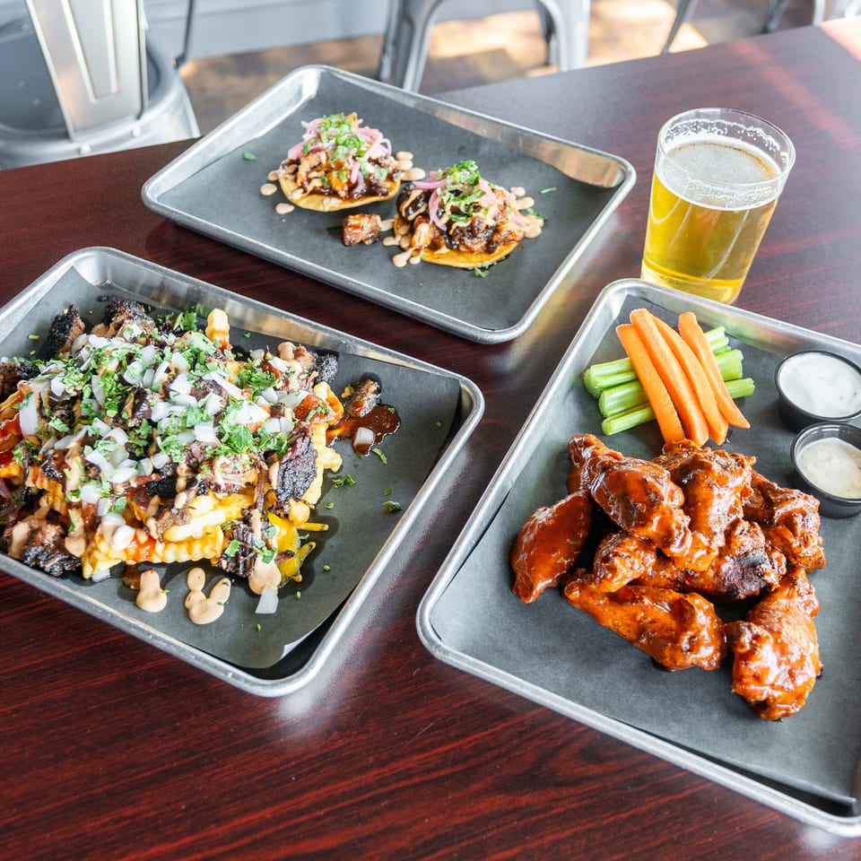 Three metal trays of food: loaded fries, two tacos, and buffalo wings with carrots, celery, and dips.