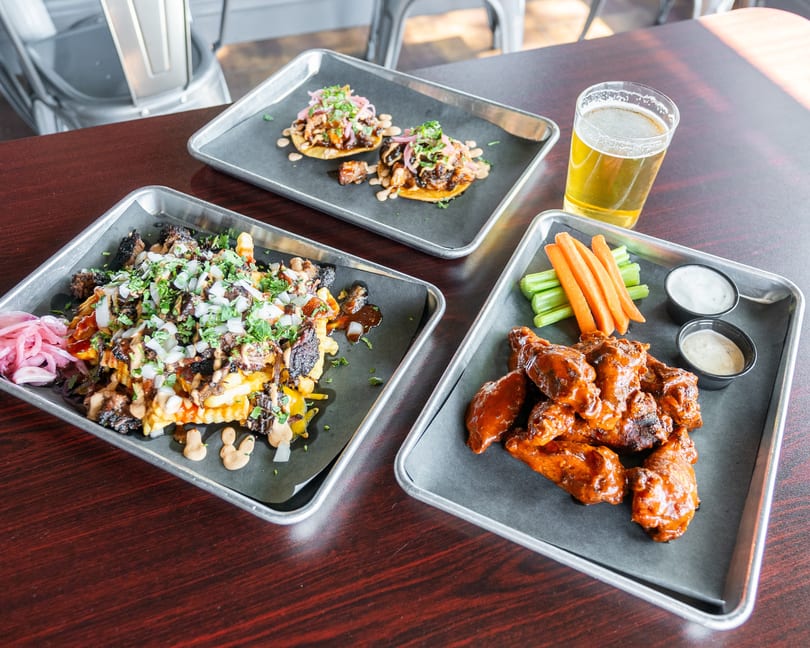 Three metal trays of food: loaded fries, two tacos, and buffalo wings with carrots, celery, and dips.