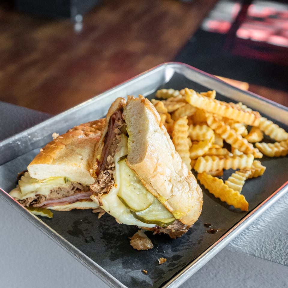 Cuban sandwich with pickles and melted cheese, served with crinkle-cut fries and a beer.