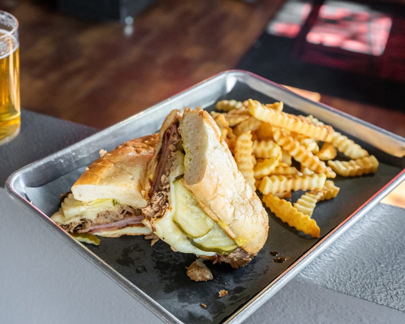 Cuban sandwich with pickles and melted cheese, served with crinkle-cut fries and a beer.