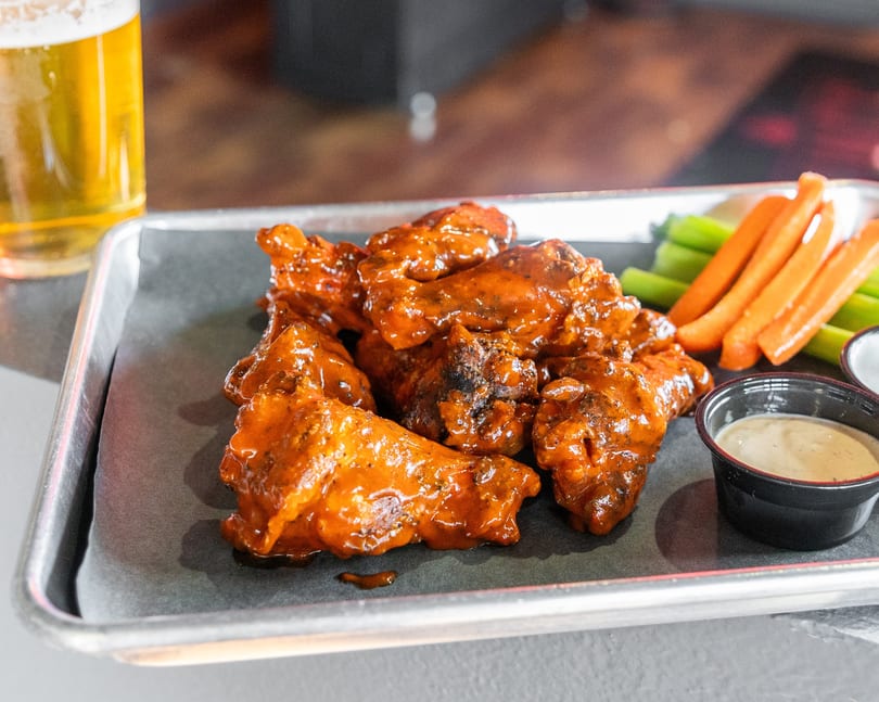 A tray of buffalo wings with celery, carrots, and dipping sauces, next to a glass of beer.
