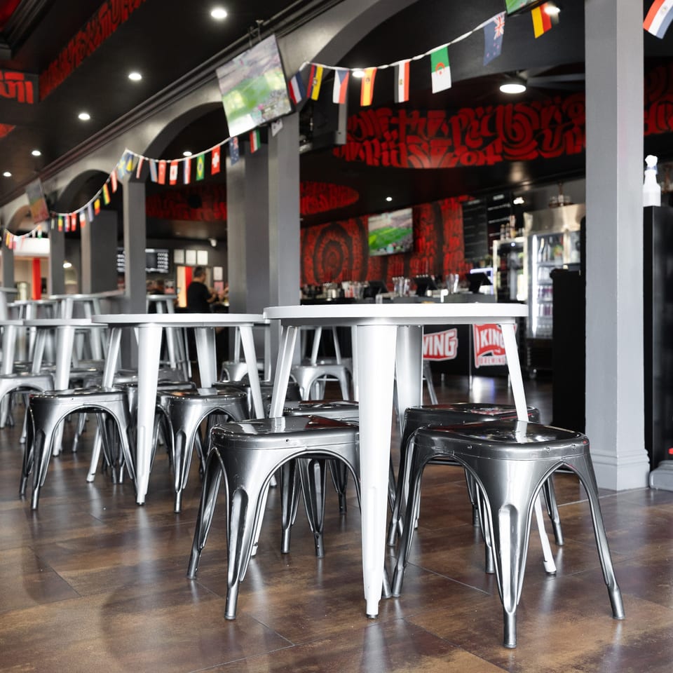Empty restaurant interior with rows of silver stools and white high-top tables, decorated with international flags.