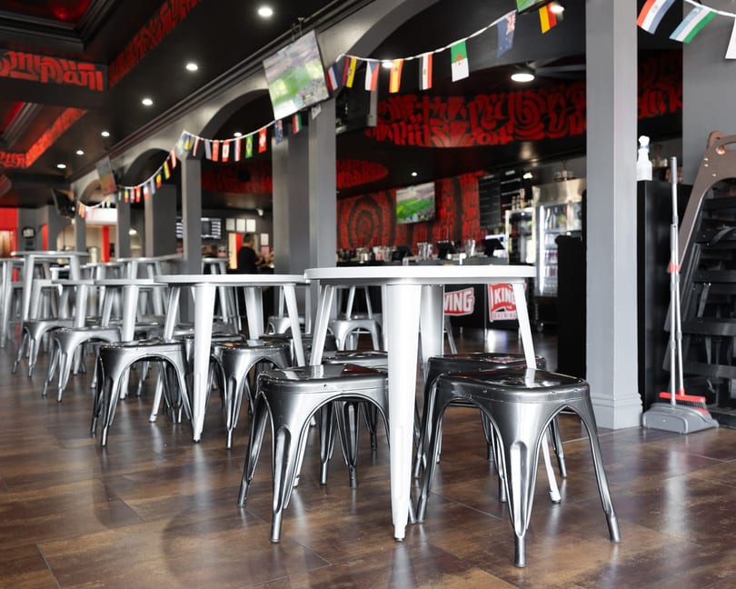 Empty restaurant interior with rows of silver stools and white high-top tables, decorated with international flags.