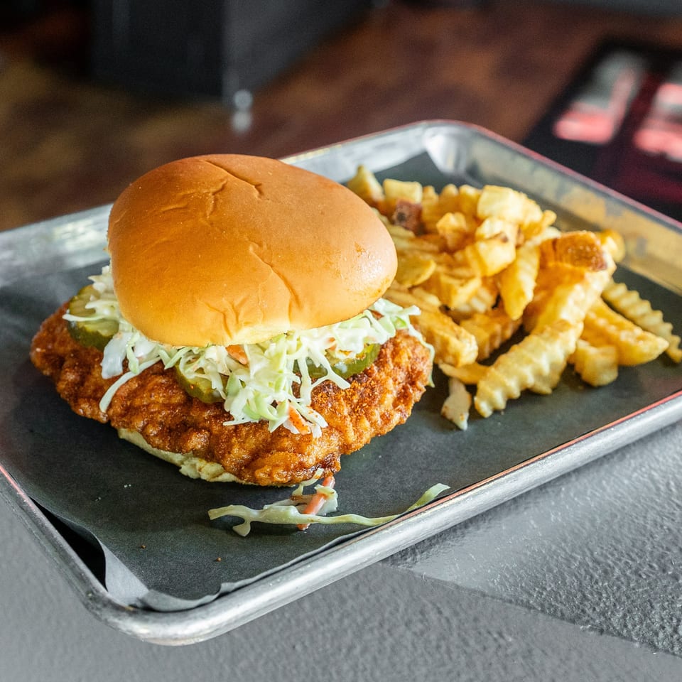 Crispy fried chicken sandwich with coleslaw and pickles, served with crinkle-cut fries on a metal tray.