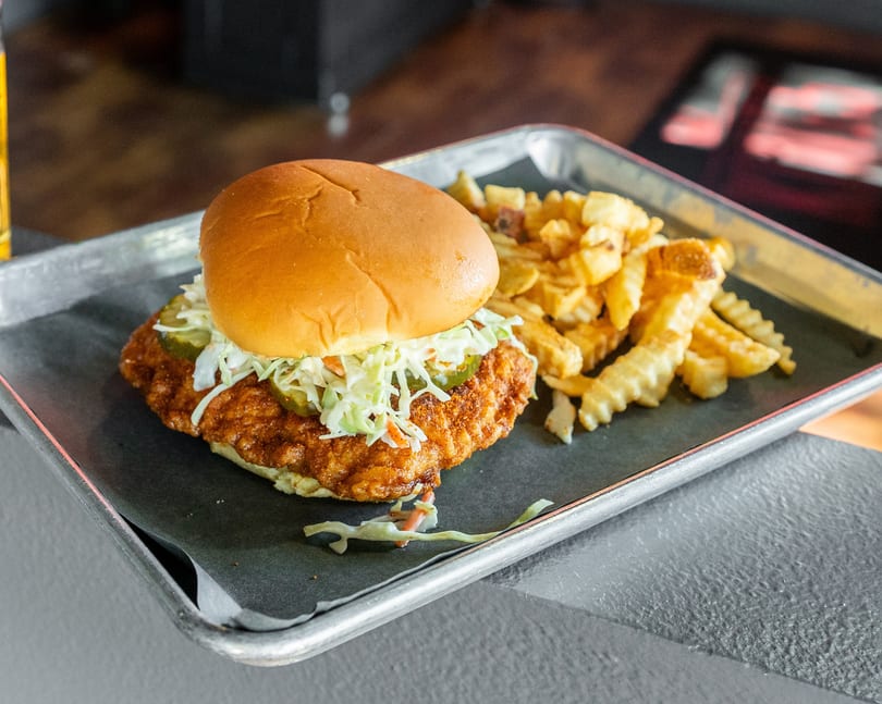 Crispy fried chicken sandwich with coleslaw and pickles, served with crinkle-cut fries on a metal tray.