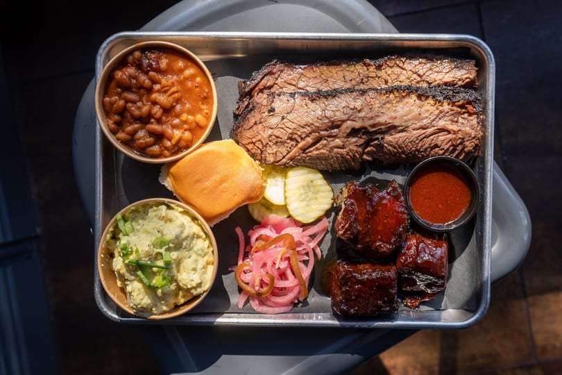 BBQ platter on a metal tray with sliced brisket, baked beans, potato salad, burnt ends, pickles, and a bun.