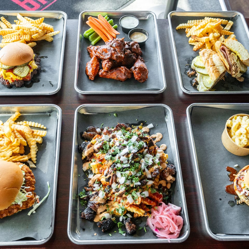Six metal trays of various BBQ dishes, including burgers, wings, sandwiches, and loaded fries.