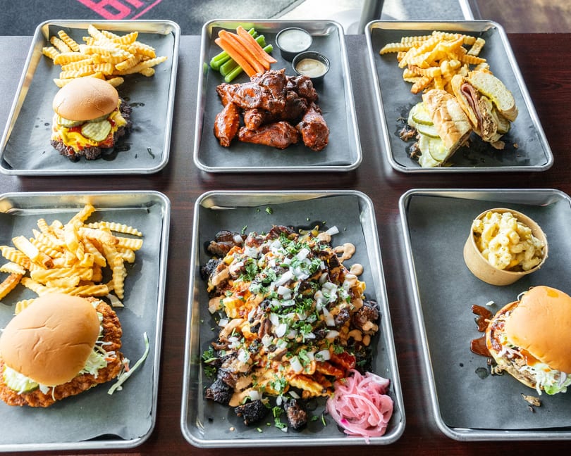 Six metal trays of various BBQ dishes, including burgers, wings, sandwiches, and loaded fries.