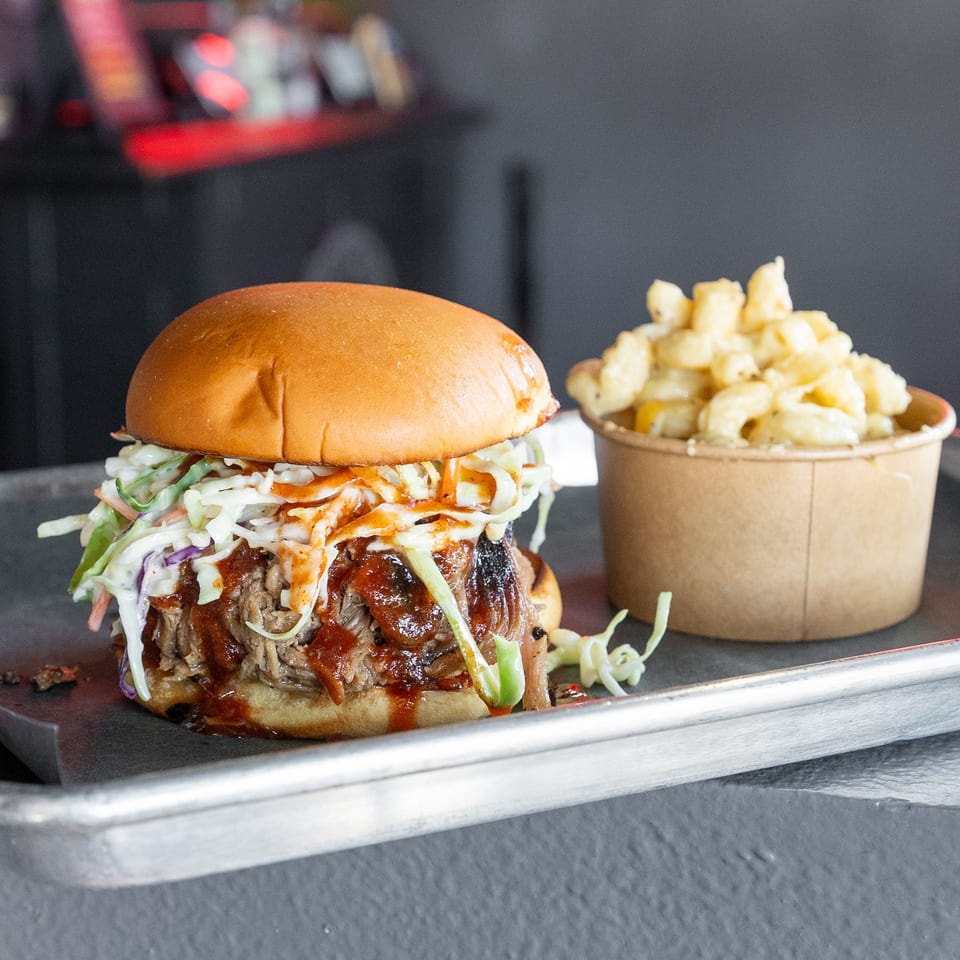Pulled pork sandwich with coleslaw and BBQ sauce, next to a cup of macaroni and cheese.