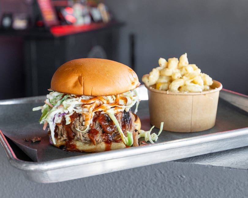 Pulled pork sandwich with coleslaw and BBQ sauce, next to a cup of macaroni and cheese.