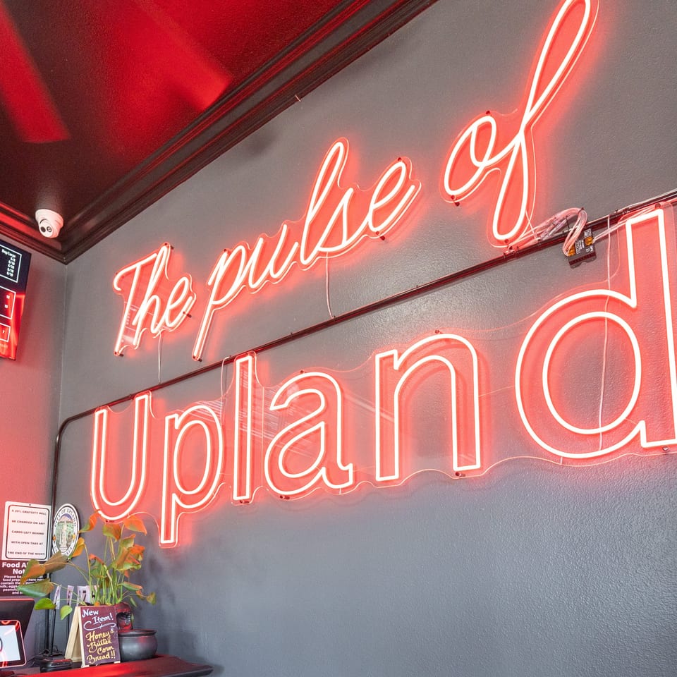 A red neon sign on a gray wall reads "The pulse of Upland" above a counter with a plant.