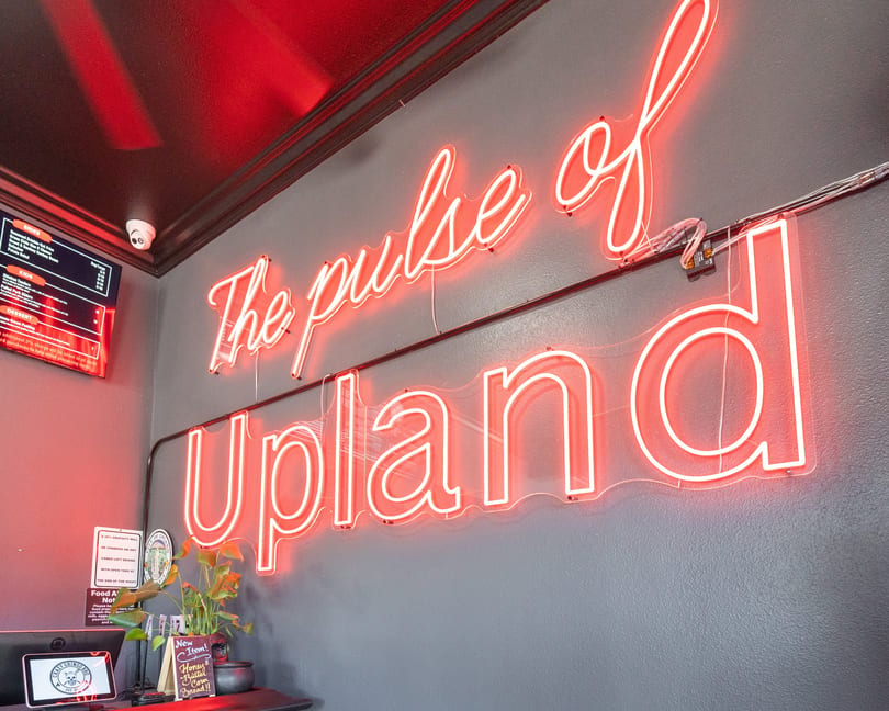 A red neon sign on a gray wall reads "The pulse of Upland" above a counter with a plant.