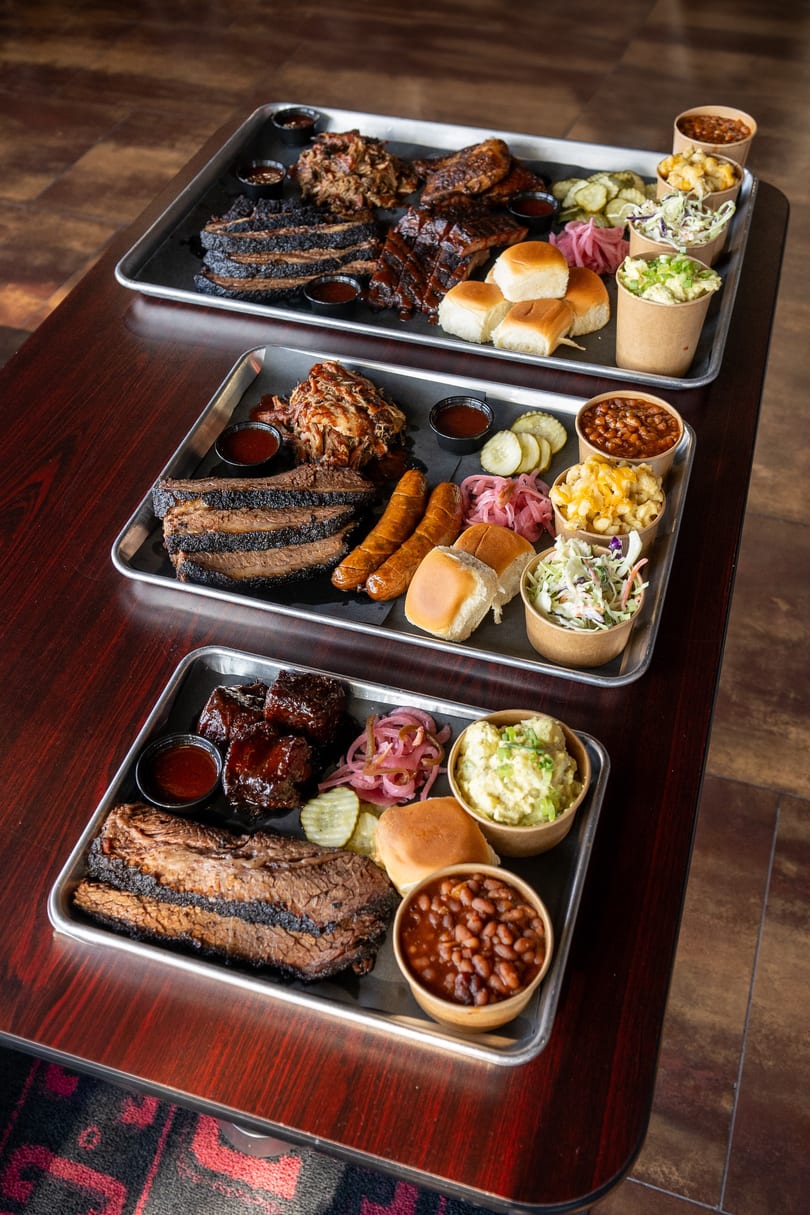 Three large BBQ platters featuring brisket, pulled pork, ribs, sausages, rolls, and assorted sides.