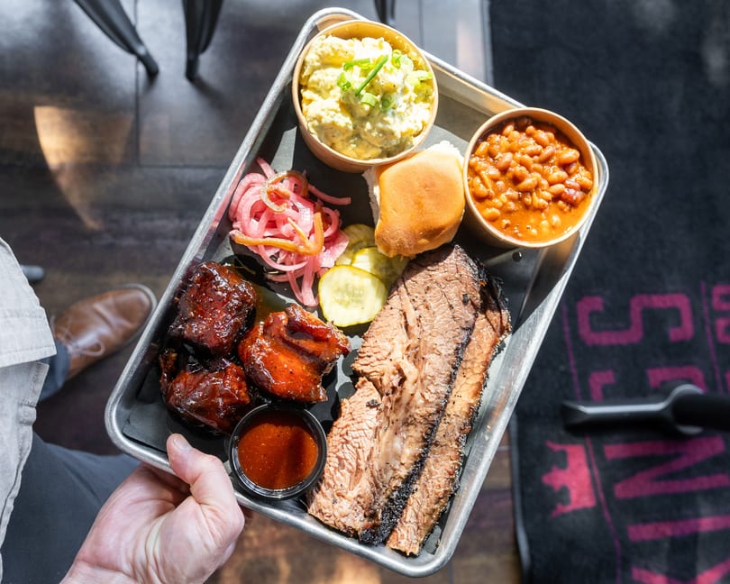 A hand holds a metal tray with brisket, ribs, baked beans, potato salad, pickles, onions, and a roll.