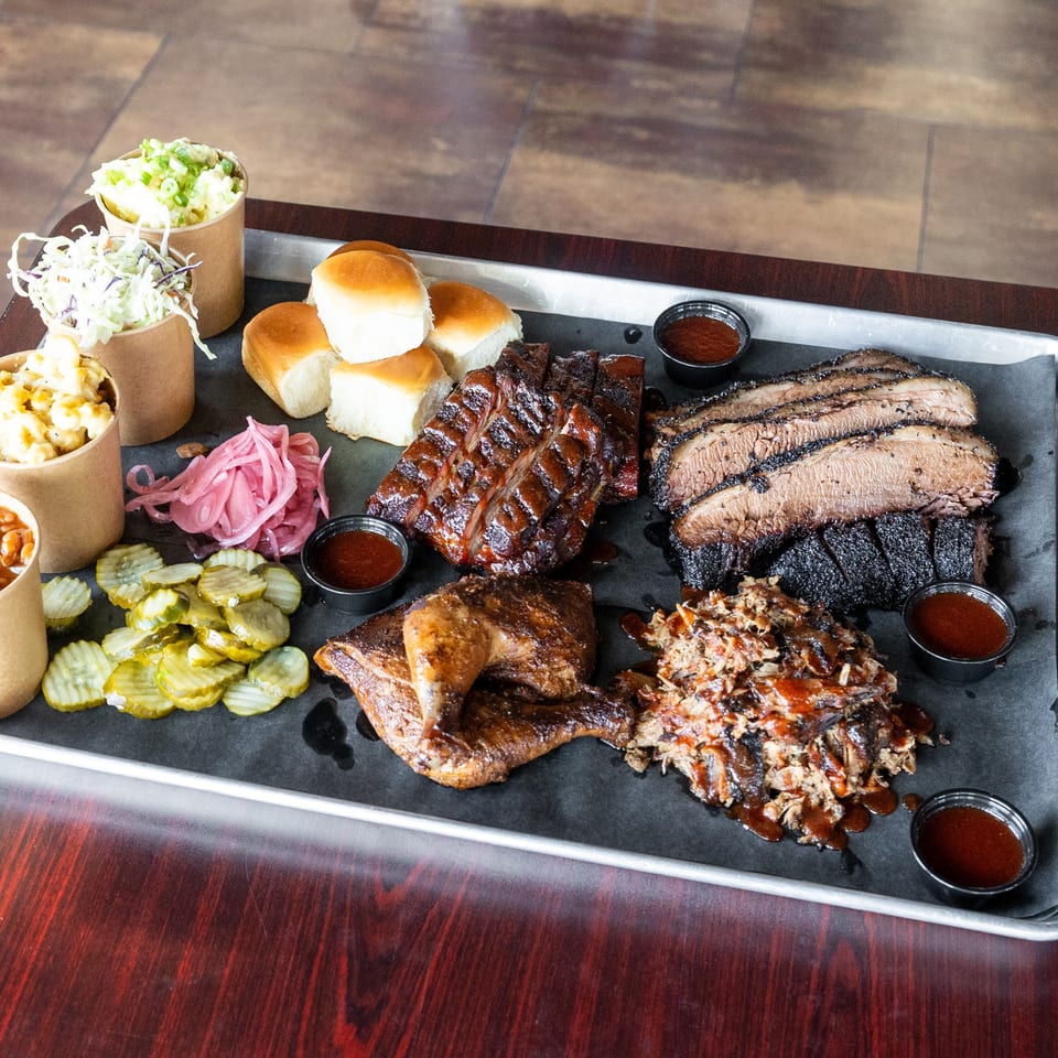 A large metal tray filled with barbecue meats, sides, and sauces on a dark wooden table.