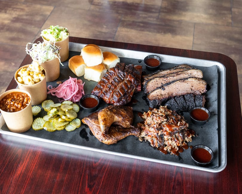 A large metal tray filled with barbecue meats, sides, and sauces on a dark wooden table.